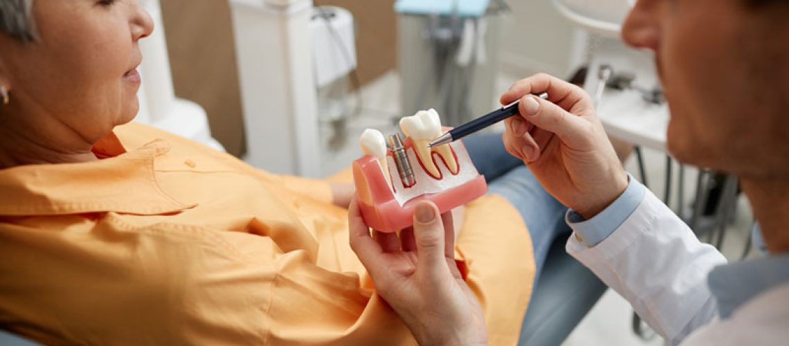 Closeup of male dentist holding tooth model while explaining dental implant surgery to a new patient in a dental clinic.